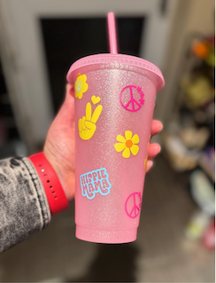 Pink glitter cup with pink, blue and yellow sticker graphics of peace signs, flowers, hearts and 'hippie mama' text.