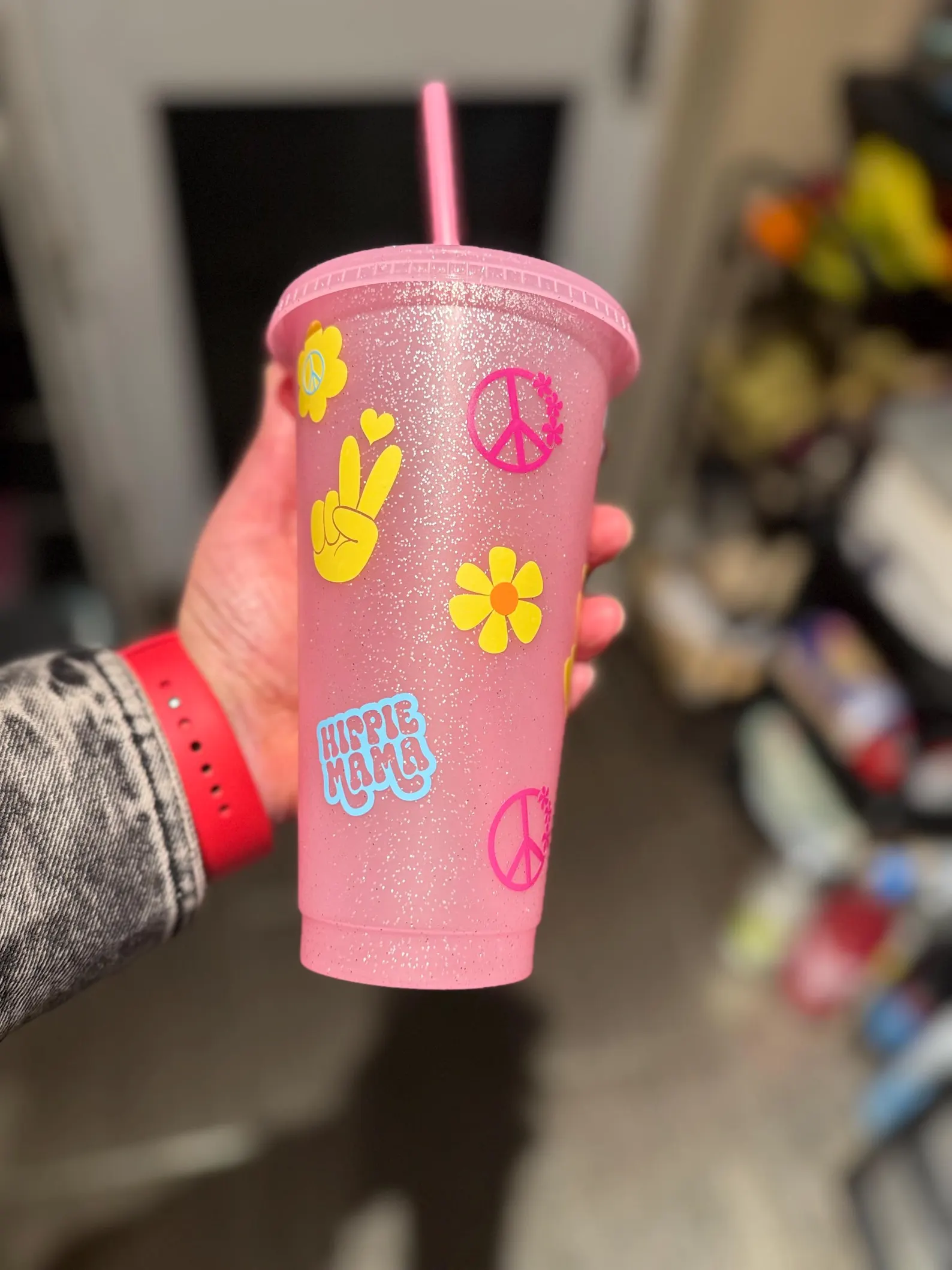 Pink glitter cup with pink, blue and yellow sticker graphics of peace signs, flowers, hearts and 'hippie mama' text.